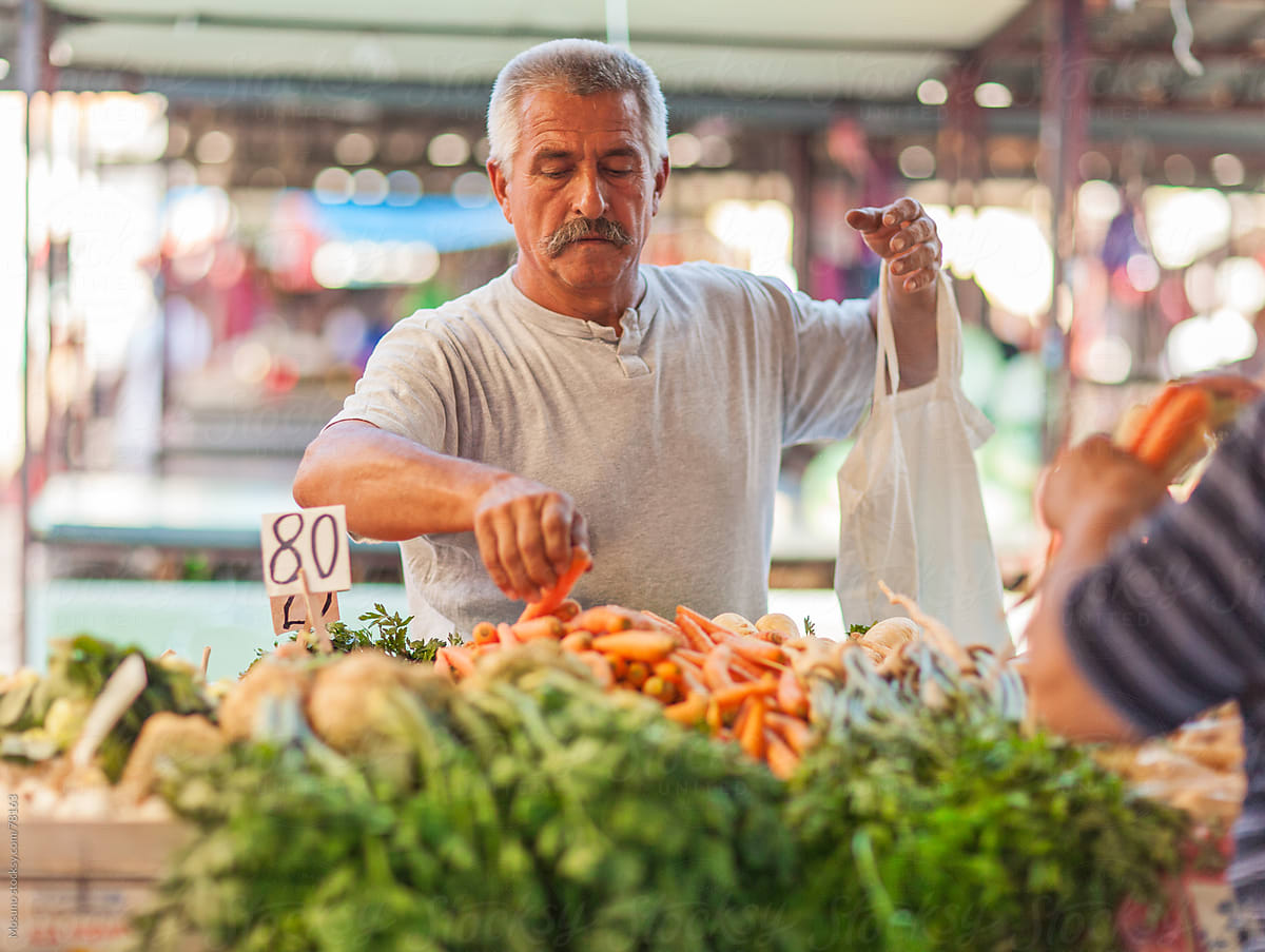 An older white man with a grey moustache, in light grey shirt, buys carrots at an open-air farmers market.