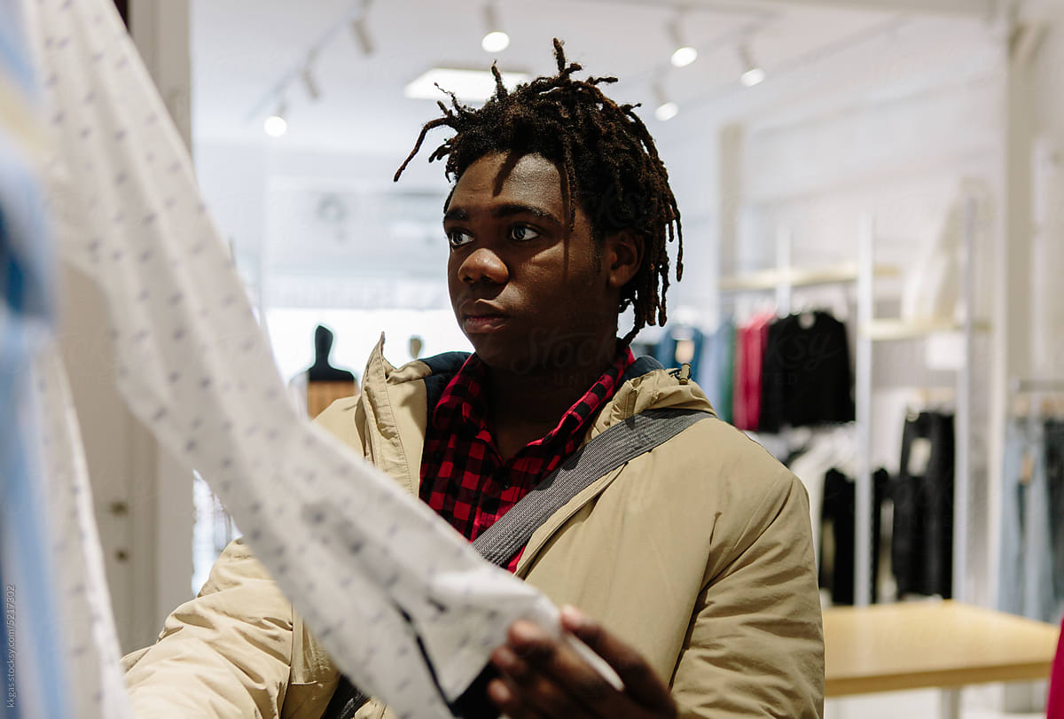 A male youth in a brightly lit modern clothing store, stares in dumb horror, as he attempts to figure out whether the long-sleeved shirt he is holding is for him or possibly women’s clothes.